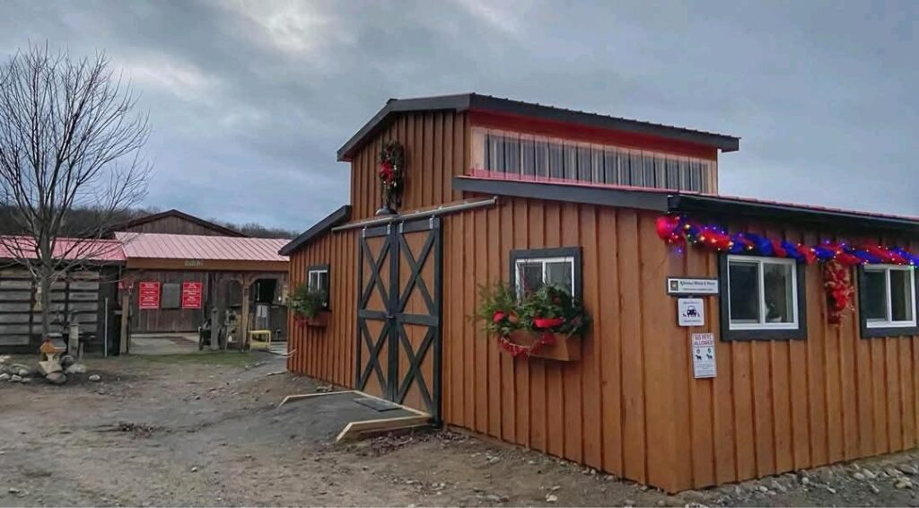 Rustic Exterior of the custom horse barn taken from outside of the barn. The siding is brown wood with some black painted x shaped accents on the closed sliding door. The end windows have greenery planters with red bows attached to the exterior of the building under the window