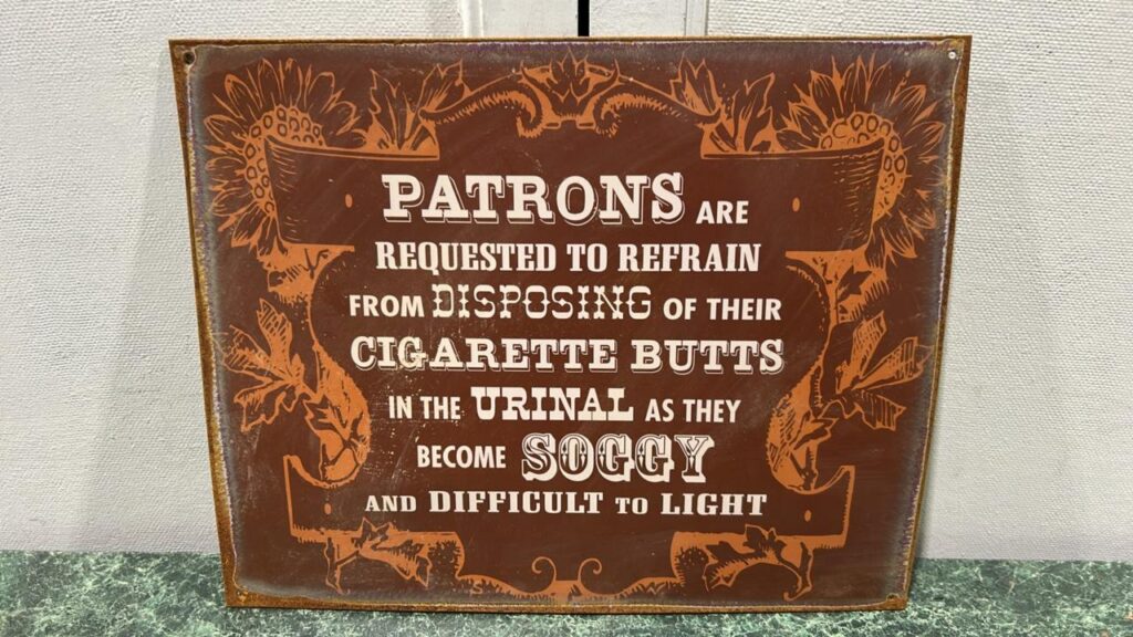 Burnt red sign that has orange coloured mosaic around the following words - Patrons - pleaserefrain from disposing of you cigarette butts in the urinal as they become soggy and difficult to light