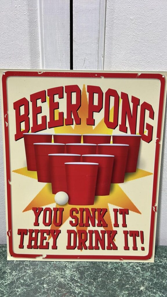 Metal sign that has a beige background with 4 rows of red cups- back row 4 cups, row in front of it is 3 cups, 2 cups in front of it and 1 cup with a ping pong ball to the left of the cup. The words above it read Beer Pong and below it read You sink it, they drink it
