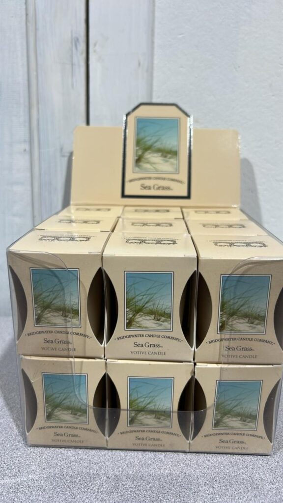 White display box containing 18 individually packaged Sea Grass Votives, Each individual box has a picture of a grass growing on a sandy hill beside the water