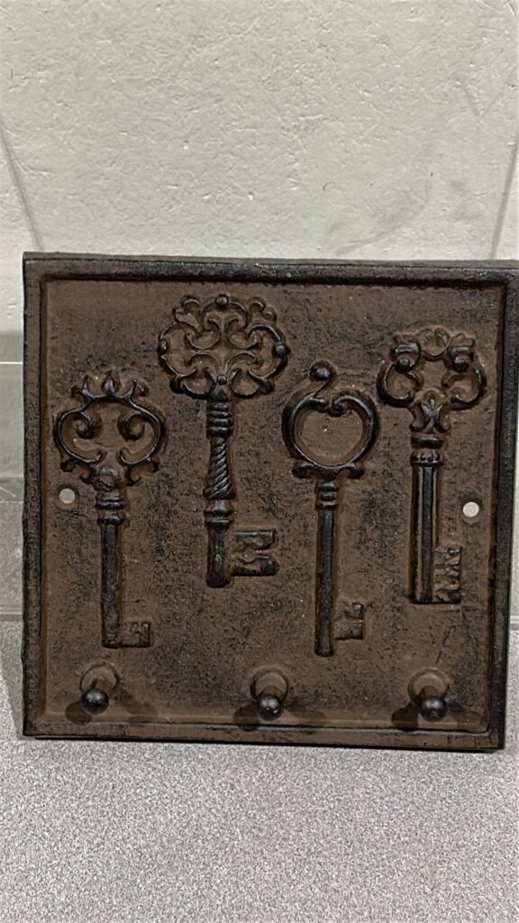 Square shaped Cast iron plaque that has 4 Vintage door keys embossed on the plaque in the middle of the rectangle. 3 hooks for keys spaced out along the bottom edge
